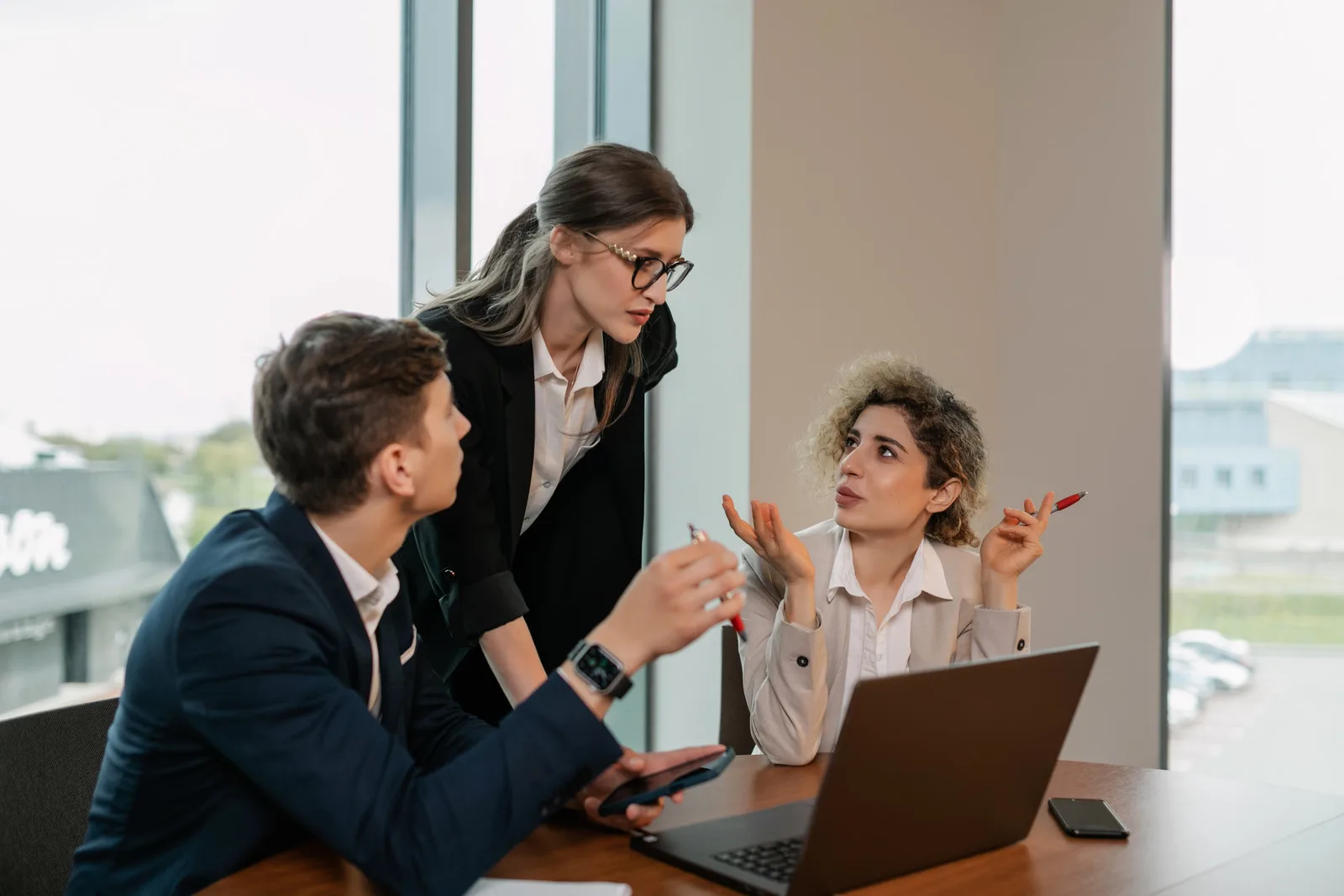 Three professionals in a business meeting, discussing work around a laptop in a modern office.